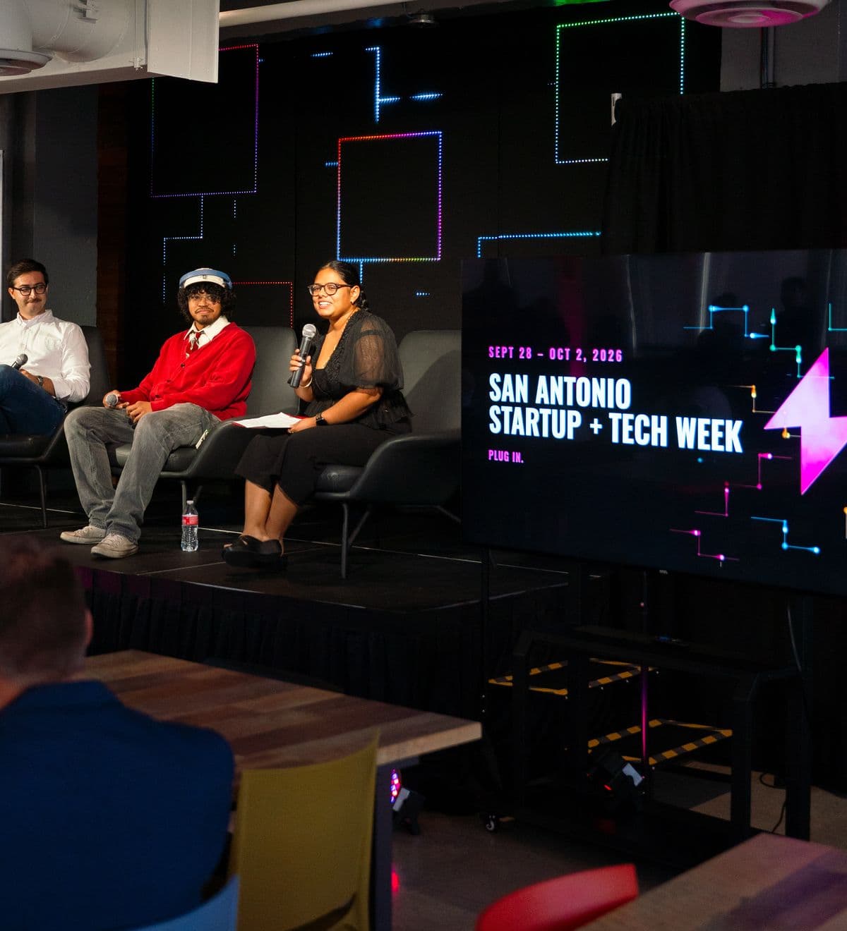 A panel on the Geekdom stage at The Rand, beside a screen reading San Antonio Startup + Tech Week, Plug in.